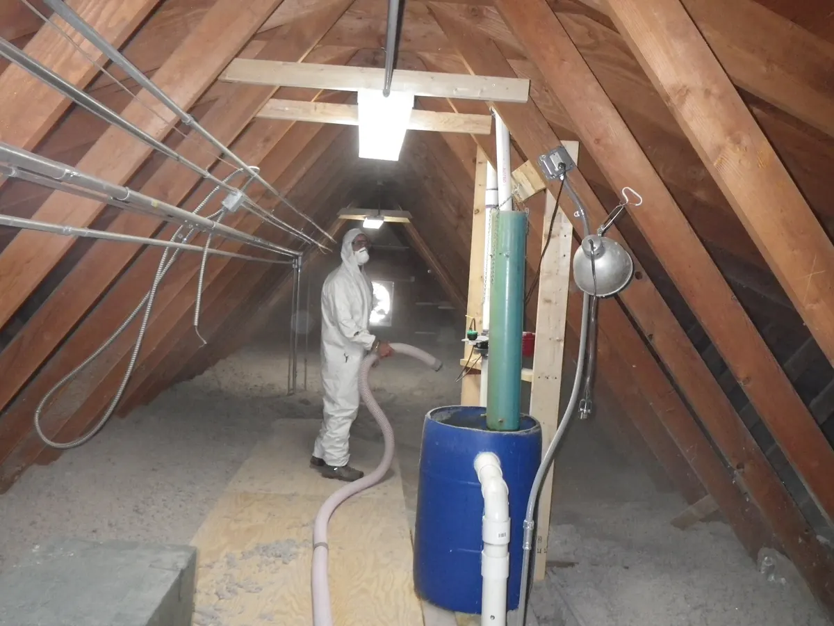 A person in coveralls and a respirator blowing loose-fill cellulose into an attic through a large hose
