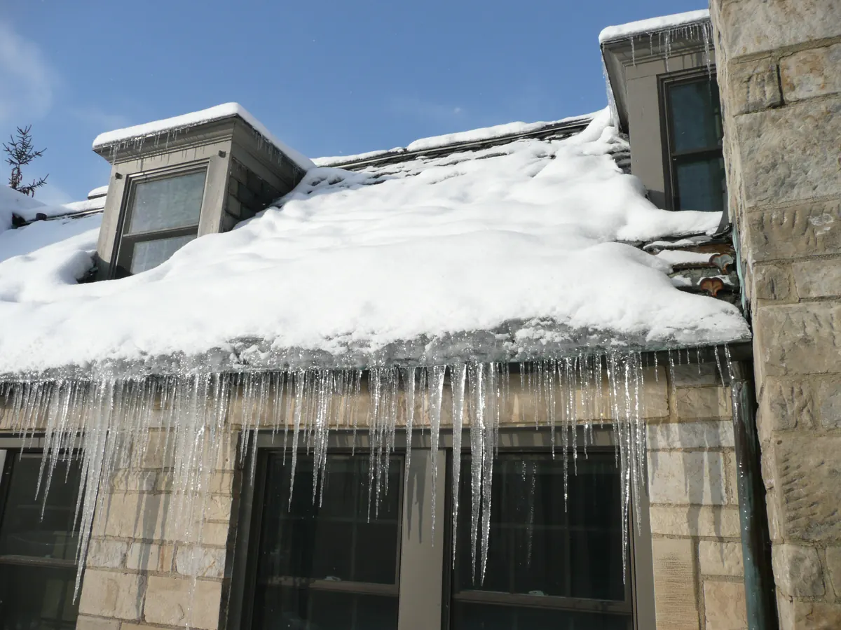 Snow-covered roof with a thick band of ice at the eave and long icicles hanging from the gutter line