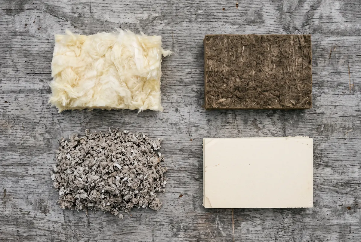 Four loose samples of building insulation laid out on plywood: fluffy yellow fiberglass, shredded gray cellulose, a dense brown mineral wool square and a cream rigid foam offcut